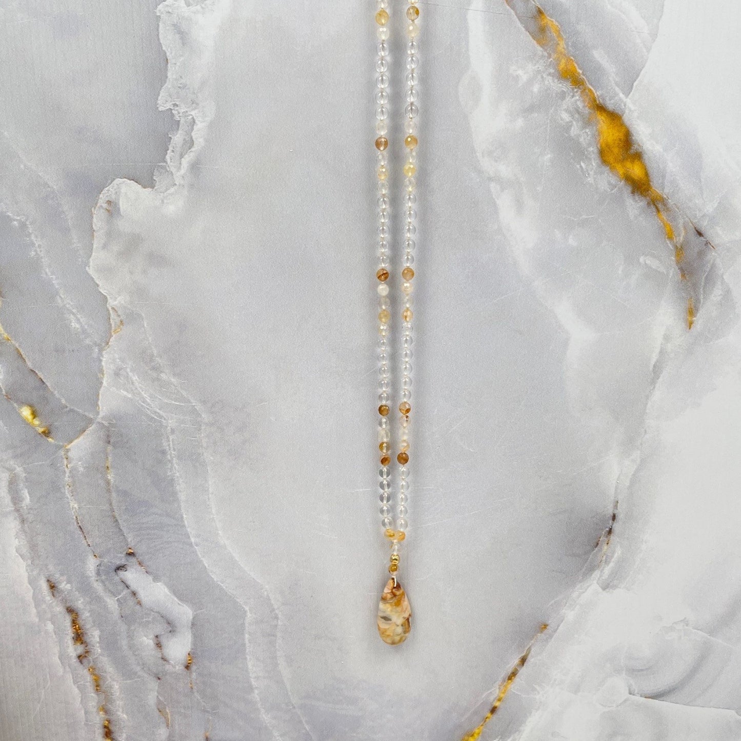 Mala ketting van bergkristal en golden healer kralen met een hanger van een gold filled kraal en een druppelvormige agaat in gele en bruine kleuren. Hier hangt de ketting voor een marmeren achterwand.